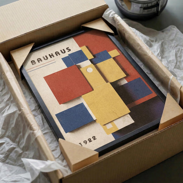 May include: Framed Bauhaus exhibition poster from 1922 featuring geometric rectangular shapes in red, blue, and yellow arranged in an abstract composition. The vintage-style poster displays "BAUHAUS" and "1922" text on cream background. Shown in black frame inside cardboard shipping box with protective tissue paper wrapping on wooden surface.