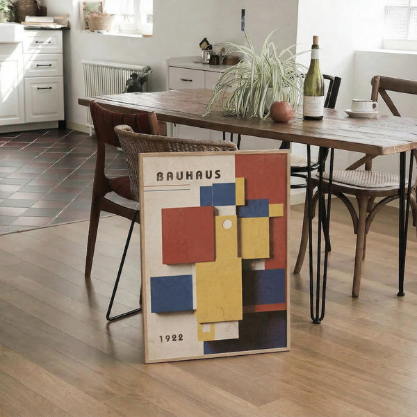 May include: Geometric abstract art poster with colorful rectangular shapes in red, blue, yellow, and orange displayed on wooden floor against dining table in modern kitchen. Poster shows Bauhaus typography and 1922 date on cream background. Room features wooden table with hairpin legs, woven dining chairs, white kitchen cabinets, and natural lighting from windows.