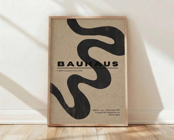 May include: Framed Bauhaus poster with flowing black serpentine shape on beige textured background, displayed against white wall on light wooden floor. Natural window light creates soft shadows. Minimalist Scandinavian-style interior setting with clean lines and neutral tones.