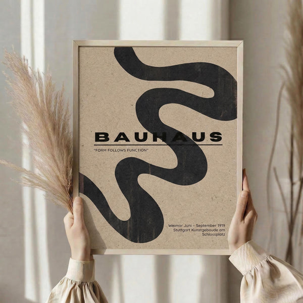 May include: Hands holding a framed beige poster with flowing black curved shapes resembling ribbons or smoke. The poster displays "BAUHAUS" text with "FORM FOLLOWS FUNCTION" subtitle and dates "Weimar Juni - September 1919 Stuttgart Kunstgeboude am Schlossplatz." Person wears cream-colored clothing with gathered sleeves, holding dried pampas grass stems. Light wooden frame against soft neutral background with natural window lighting.