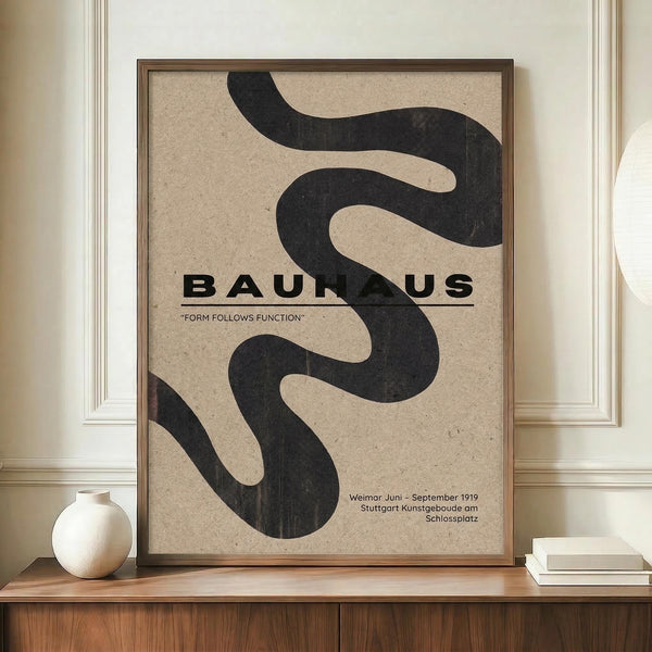 May include: Framed Bauhaus poster with flowing black abstract shapes on beige textured background, displayed on wooden sideboard next to white ceramic vase and stacked books in elegant room with white wainscoting and cream walls. Poster features bold typography reading "BAUHAUS" and "FORM FOLLOWS FUNCTION" with exhibition details for Weimar and Stuttgart locations dated 1919.