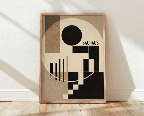 May include: Framed Bauhaus-style geometric abstract poster featuring black circle, rectangles, and vertical lines arranged on cream and brown background, displayed in light wood frame leaning against white wall on natural wood flooring with soft natural lighting creating shadows.