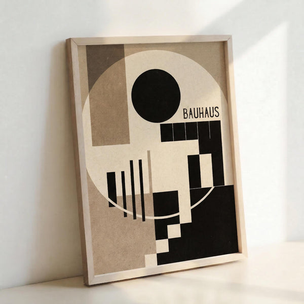 A framed geometric abstract poster featuring Bauhaus-style design with black and beige shapes, including a large white circle, black circle, vertical lines, and rectangular forms arranged in a modernist composition. The word "BAUHAUS" appears in black text. The artwork is displayed in a light wood frame leaning against a white wall.