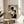 May include: Framed geometric abstract poster in beige, black and white featuring circles, rectangles and Bauhaus text, displayed on light wood round side table next to beige upholstered chair with white throw pillow. Natural sunlight streams across neutral-toned room with plant leaves visible in corner. Thin natural wood frame complements minimalist Scandinavian-style furniture and decor.