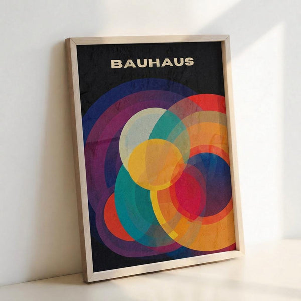 Framed poster featuring overlapping translucent circles in vibrant colors including yellow, orange, red, blue, purple, and teal against a black background. The word "BAUHAUS" appears in cream letters at the top. The artwork has a geometric, modernist style with layered circular forms creating color blending effects where they intersect. Displayed in a light wooden frame, leaning against a white wall.