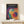 May include: Framed Bauhaus-style poster with overlapping colorful circles in blues, purples, reds, yellows, and greens on dark background, displayed on white shelf next to small wrapped gift with natural twine and greenery sprig against cream wall