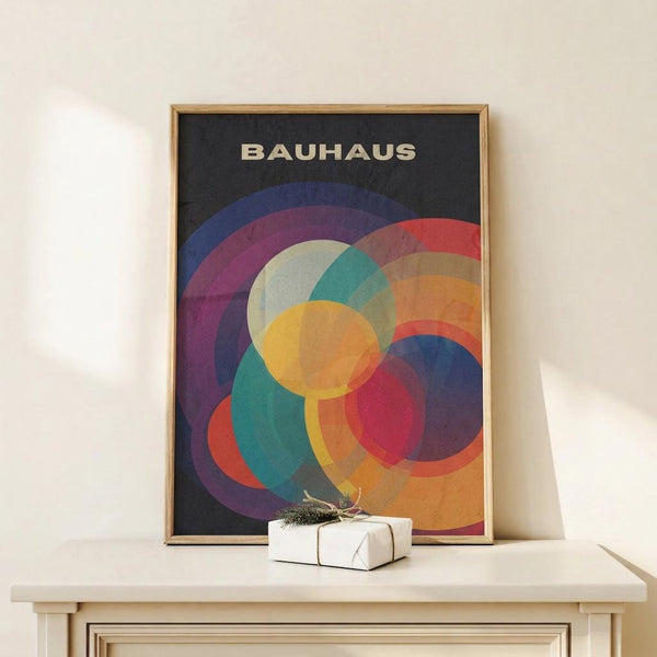 May include: Framed Bauhaus-style poster with overlapping colorful circles in blues, purples, reds, yellows, and greens on dark background, displayed on white shelf next to small wrapped gift with natural twine and greenery sprig against cream wall