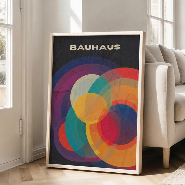 May include: Framed poster with overlapping colorful circles in vibrant blues, purples, reds, oranges, yellows, and teals on black background with "BAUHAUS" text at top, displayed in light wood frame leaning against white wall next to beige upholstered chair in bright living room with large windows and wooden floors.