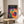 May include: Colorful geometric poster with overlapping translucent circles in vibrant blues, purples, reds, oranges, yellows, and teals against black background, displayed in thin wooden frame on round natural wood side table next to white upholstered chair, with plant leaves visible and soft natural lighting creating shadows on white textured wall behind.