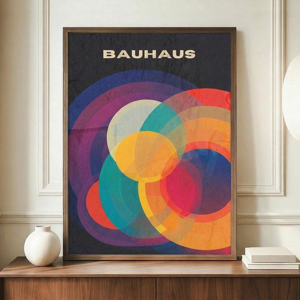 May include: Framed Bauhaus-style poster with overlapping geometric circles in vibrant colors including purple, orange, teal, red, and yellow on dark background, displayed on wooden credenza next to white ceramic vase and stacked books in elegant room with white wainscoting.
