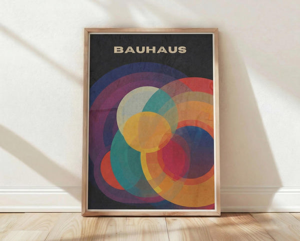 May include: Framed Bauhaus-style poster with overlapping translucent circles in vibrant colors including blue, purple, red, orange, yellow, and teal on dark background. Light wood frame leaning against white wall on light wooden floor with natural window shadows.