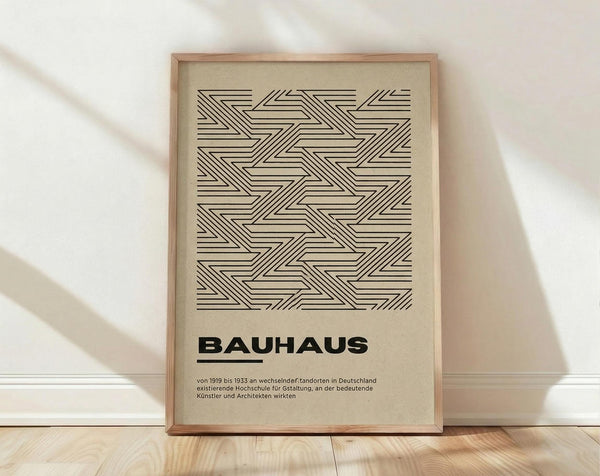 May include: Framed geometric poster with black linear zigzag pattern on cream background, displaying "BAUHAUS" text and German description below. Light wood frame leaning against white wall corner on natural wood flooring with soft natural lighting and shadows.
