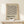May include: Framed beige poster with black geometric zigzag line pattern and "BAUHAUS" text in bold letters, displayed on light wood shelf against cream wall. Small wrapped white gift box with twine and evergreen sprig sits in front of the thin wooden frame.