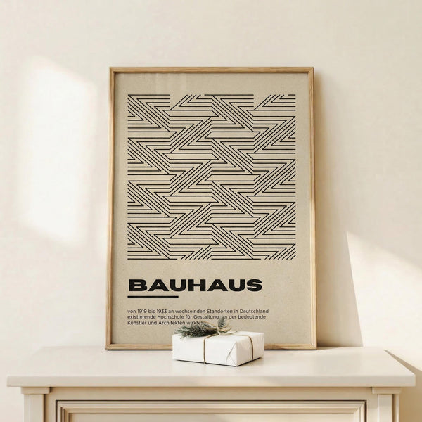 May include: Framed beige poster with black geometric zigzag line pattern and "BAUHAUS" text in bold letters, displayed on light wood shelf against cream wall. Small wrapped white gift box with twine and evergreen sprig sits in front of the thin wooden frame.
