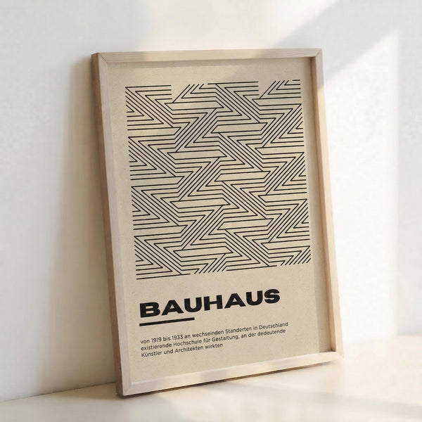 May include: Framed geometric poster with black linear zigzag pattern on cream background, displayed in light wooden frame leaning against white wall. Bold "BAUHAUS" text below intricate interlocking angular design, with small German descriptive text underneath. Clean minimalist styling with natural lighting creating soft shadows.