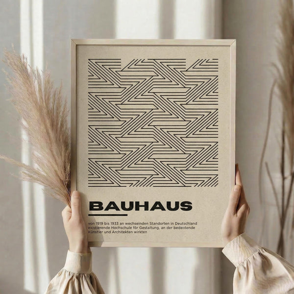May include: Person holding a framed geometric art print featuring black linear zigzag and chevron patterns on beige background, displayed in natural wood frame. Dried pampas grass visible to left. Person wears cream colored long-sleeve top with gathered cuffs. Soft natural lighting against neutral curtained background creates minimalist aesthetic.