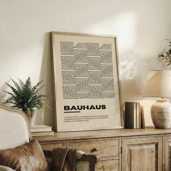 May include: Framed geometric art print with black linear pattern on cream background, displayed on rustic wooden sideboard. Scene shows neutral living room with beige armchair, potted fern, ceramic vase, and stacked books. Natural lighting creates warm shadows on white wall. Print features interlocking angular lines forming repeating tessellated design with German text below pattern.