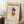 May include: Framed Bauhaus poster with geometric design featuring overlapping blue and red diamond shapes on cream background, leaning against white wall next to beige armchair and wooden side table with white coffee cup. Light wood frame matches minimalist room setting with white shag rug.