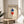 May include: Framed Bauhaus poster with geometric diamond shapes in blue and red on cream background, displayed on round wooden side table next to beige upholstered chair. Natural lighting creates soft shadows on white wall. Green plant leaves visible in corner. Modern minimalist room setting showcases the vintage-style geometric art print in thin wooden frame.