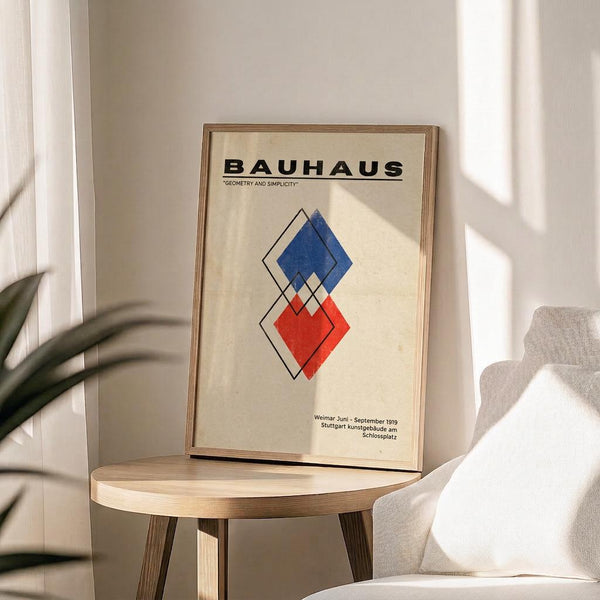 May include: Framed Bauhaus poster with geometric diamond shapes in blue and red on cream background, displayed on round wooden side table next to beige upholstered chair. Natural lighting creates soft shadows on white wall. Green plant leaves visible in corner. Modern minimalist room setting showcases the vintage-style geometric art print in thin wooden frame.