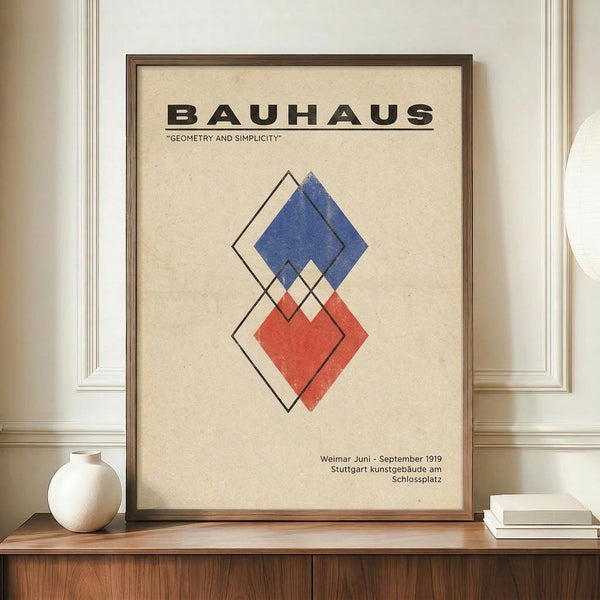 May include: Framed Bauhaus poster with geometric diamond shapes in blue and red colors, displayed on wooden credenza next to white ceramic vase and stacked books against cream paneled wall. Poster features vintage-style design with overlapping angular forms and German text referencing Weimar 1919 exhibition.