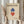 May include: Hands holding a framed Bauhaus-style poster featuring geometric diamond shapes in blue and red with black outlines on beige background. Person wears cream colored clothing and holds dried wheat stems. Neutral curtained interior setting with natural lighting.