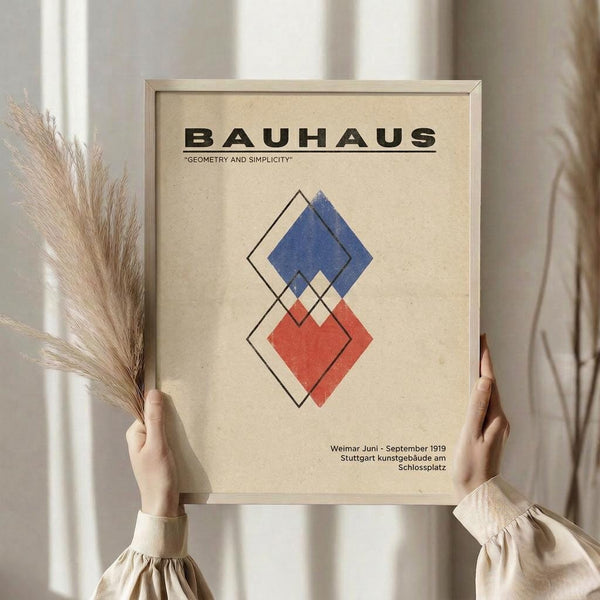 May include: Hands holding a framed Bauhaus-style poster featuring geometric diamond shapes in blue and red with black outlines on beige background. Person wears cream colored clothing and holds dried wheat stems. Neutral curtained interior setting with natural lighting.