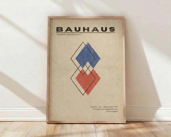 May include: Framed Bauhaus-style poster with geometric diamond shapes in blue and red on cream background, displayed against white wall on light wood floor. Features vintage typography and minimalist design aesthetic typical of 1919 Weimar art movement.