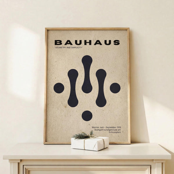 May include: Framed Bauhaus poster with beige textured background displaying black geometric shapes - circles and elongated forms arranged symmetrically. Bold black text reads "BAUHAUS" and "GEOMETRY AND SIMPLICITY" at top, with German exhibition details at bottom. Natural wood frame sits on light wooden shelf against white wall, with small white wrapped gift beside it.