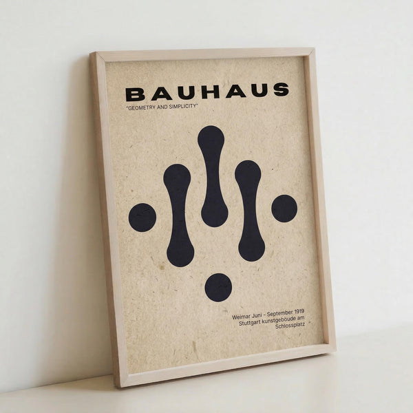 May include: Framed Bauhaus-style poster with geometric black shapes on textured beige background, displayed in light wooden frame leaning against white wall. Design features abstract droplet and circle forms arranged symmetrically with "BAUHAUS" and "GEOMETRY AND SIMPLICITY" text at top, exhibition details at bottom in minimalist typography.