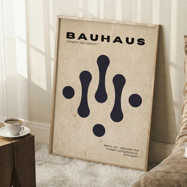 May include: Framed Bauhaus poster with cream textured background and black geometric shapes arranged vertically - circles and elongated teardrop forms. Black text reads "BAUHAUS" and "GEOMETRY AND SIMPLICITY" at top, with German text at bottom referencing Weimar and Stuttgart locations. Light wood frame, displayed leaning against white wall corner on textured rug, with coffee cup on wooden block nearby.