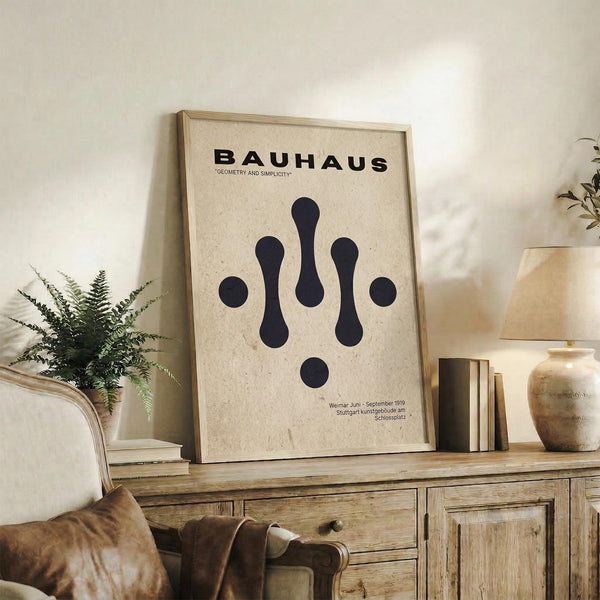 May include: Framed Bauhaus poster with geometric black shapes on cream background, displayed on rustic wooden sideboard alongside fern plant, ceramic vase, and books in neutral-toned living room setting with beige chair and soft lighting