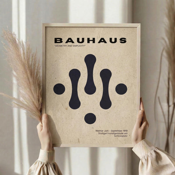 May include: Person in beige clothing holding framed Bauhaus poster with geometric black shapes on tan textured background, dried pampas grass visible to left, soft natural lighting from window, minimalist neutral interior setting