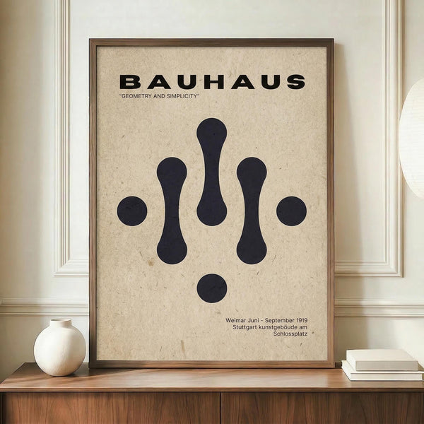 May include: Framed Bauhaus-style poster with beige textured background displaying black geometric shapes - circles and elongated oval forms arranged symmetrically. Bold black "BAUHAUS" text at top with "GEOMETRY AND SIMPLICITY" subtitle. German exhibition text at bottom. Wooden frame sits on warm wood sideboard beside white ceramic vase and stacked books against cream paneled wall.
