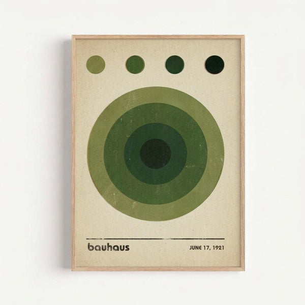 A framed poster with geometric green circles in varying shades from light olive to dark forest green arranged concentrically on cream paper. Four small circles at the top progress from light to dark green. Text at bottom reads "bauhaus" and "JUNE 17, 1921" in simple black font. The artwork has a minimalist, mid-century modern aesthetic with clean lines and is displayed in a natural wood frame against a white wall.