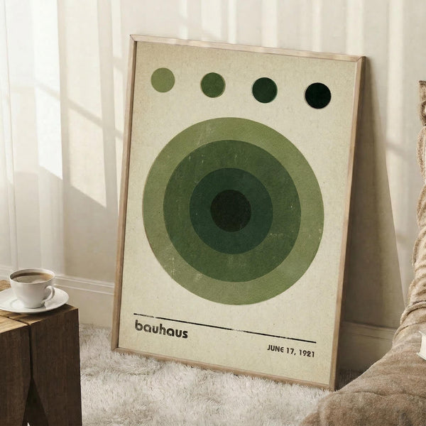 May include: Framed Bauhaus-style poster with concentric green circles in varying shades from light to dark, four small circles across the top, and "bauhaus" with "JUNE 17, 1921" text at bottom. Cream background in natural wood frame, leaning against white wall corner next to coffee cup on wooden block and textured throw blanket.