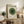 May include: Framed Bauhaus-style art print featuring concentric green circles in varying shades from light to dark green, with four small dots above, displayed on rustic wooden sideboard next to fern plant, books, ceramic vase, and leather chair in warm-toned living room setting.