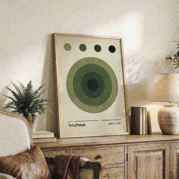 May include: Framed Bauhaus-style art print featuring concentric green circles in varying shades from light to dark green, with four small dots above, displayed on rustic wooden sideboard next to fern plant, books, ceramic vase, and leather chair in warm-toned living room setting.