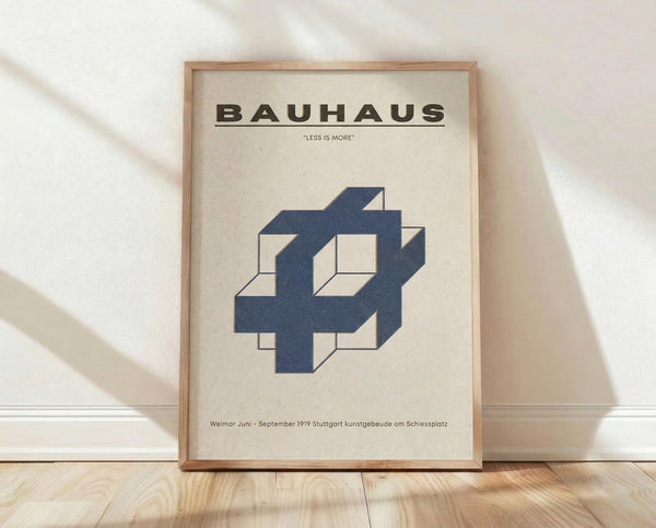 May include: Framed Bauhaus poster with minimalist blue geometric interlocking design on cream background, displayed in light wooden frame leaning against white wall on natural wood floor. Poster features "BAUHAUS" text at top, "LESS IS MORE" quote, and German text at bottom. Soft natural lighting creates diagonal shadows across the scene.