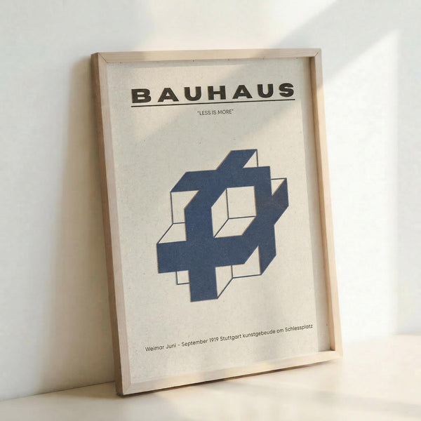May include: Framed Bauhaus poster print with cream background featuring bold black text reading "BAUHAUS" and "LESS IS MORE" above a geometric blue interlocking cube design. Text at bottom references Weimar Juni September 1919 Stuttgart kunstgebeude om Schlesplatz. Print displayed in light wood frame leaning against white wall on white surface, showing minimalist modernist design aesthetic.