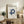 May include: White cream poster with BAUHAUS text and Less is More quote, featuring blue geometric interlocking shapes design, displayed in thin wooden frame on rustic wood sideboard alongside fern plant, stacked books, ceramic vase, and brown leather chair in modern minimalist living room setting