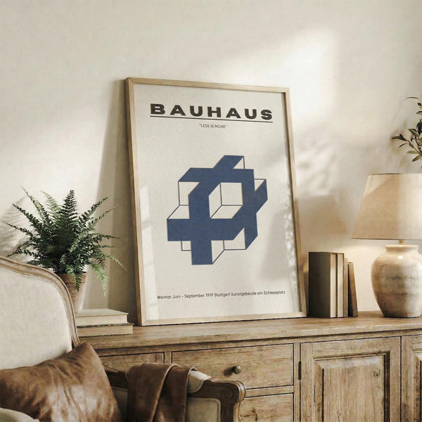 May include: White cream poster with BAUHAUS text and Less is More quote, featuring blue geometric interlocking shapes design, displayed in thin wooden frame on rustic wood sideboard alongside fern plant, stacked books, ceramic vase, and brown leather chair in modern minimalist living room setting