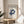 May include: Framed Bauhaus poster with "LESS IS MORE" quote and blue geometric interlocking cube design, displayed on light wood round side table next to beige upholstered chair. Natural sunlight streams across cream walls creating soft shadows, with green plant leaves visible in corner of minimalist modern interior setting.