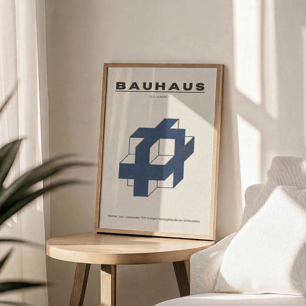 May include: Framed Bauhaus poster with "LESS IS MORE" quote and blue geometric interlocking cube design, displayed on light wood round side table next to beige upholstered chair. Natural sunlight streams across cream walls creating soft shadows, with green plant leaves visible in corner of minimalist modern interior setting.