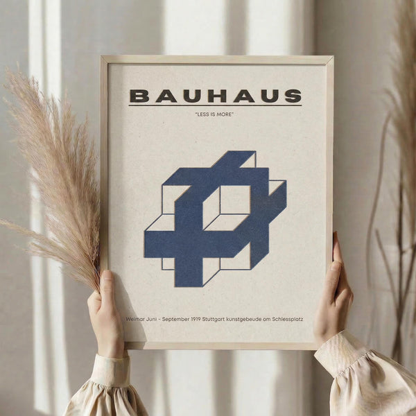 May include: Person holding framed Bauhaus poster with blue geometric interlocking shapes on cream background, wooden frame, neutral interior setting with dried pampas grass, soft natural lighting through curtains