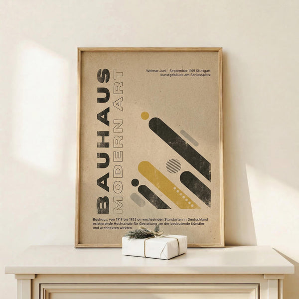 May include: Framed Bauhaus-style poster with beige background displaying "BAUHAUS MODERN ART" text and geometric shapes in black, yellow, and green. Features German text about Weimar period. Wood frame sits on light wooden surface next to small white wrapped gift with twine and greenery sprig against cream wall.