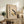 May include: Framed beige poster with "BAUHAUS MODERN ART" text and geometric shapes in yellow, black, and white displayed on rustic wooden sideboard. Setting shows brown leather chair, fern plant in white pot, ceramic vase, and books creating cozy living room scene with neutral wall background.