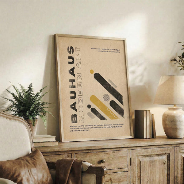 May include: Framed beige poster with "BAUHAUS MODERN ART" text and geometric shapes in yellow, black, and white displayed on rustic wooden sideboard. Setting shows brown leather chair, fern plant in white pot, ceramic vase, and books creating cozy living room scene with neutral wall background.