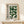 May include: Framed Bauhaus-inspired poster with green geometric semicircle pattern and "BAUHAUS MODERN ART" text in German, displayed on white wooden cabinet against cream wall. Small wrapped gift box with twine sits beside the light wood frame. Clean, minimalist styling with natural lighting creating soft shadows.
