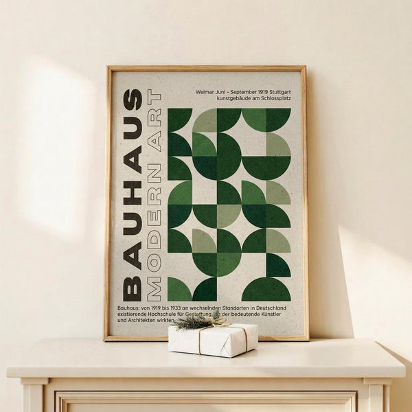May include: Framed Bauhaus-inspired poster with green geometric semicircle pattern and "BAUHAUS MODERN ART" text in German, displayed on white wooden cabinet against cream wall. Small wrapped gift box with twine sits beside the light wood frame. Clean, minimalist styling with natural lighting creating soft shadows.